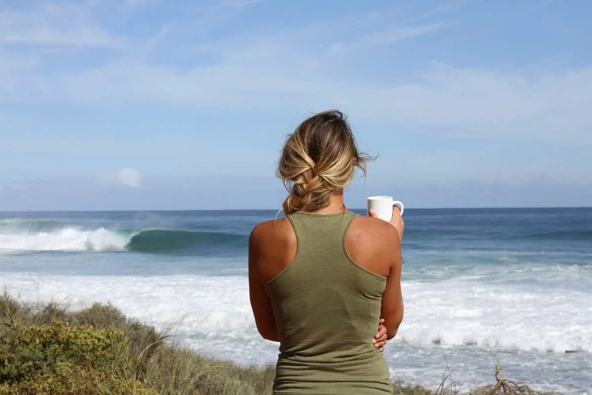 A lively woman with mushroom coffee on a beach and and reconsidering the decision of drinking it.